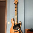 Elektrická basová kytara s přírodním dřevěným povrchem těla a hmatníkem. Na hlavě je viditelné logo Fender. Tělo má černý pickguard a čtyři ladicí ko…