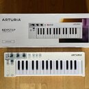 Bílá klávesová kontrola s logem ARTURIA a názvem KEYSTEP. Nástroj má plastové tělo, bílé klávesy a ovládací prvky v horní části. Vedle nástroje je um…