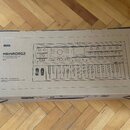 Kartonová krabice s černobílým tiskem znázornujícím syntetizátor. Na krabici jsou nápisy KORG, microKORG2, SYNTHESIZER/VOCODER, SYNTHÉTISEUR/VOCODEUR…