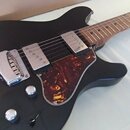 Černá elektrická kytara s hnědým hmatníkem a tmavě červeným želvovinovým pickguardem. Nástroj má dva snímače typu humbucker s kovovými kryty, chromov…