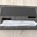 Černá papírová krabice s vložkou z šedé pěny. V dolním výřezu je umístěn předmět zabalený v bílém ochranném plastu. V horním výřezu se nachází plochý…