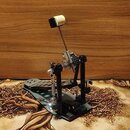 Fotografie znázorňuje mechanismus basového pedálu v černé barvě s kovovými prvky. Konstrukce obsahuje šlapadlo, řetězový převod a horní pedál s bílou…