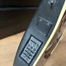 Detailní pohled na hranu černé kytary s texturovaným povrchem a krémovým bindingem. Na hraně je umístěn černý plastový kryt baterie a ovládací panel…