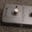 Stříbrný efekt pedál s nápisem Noise Gate a logem JOYO. Na horní straně se nachází stoupací spínač s nápisem TRUE BYPASS, bílý otočný knoflík Thresho…
