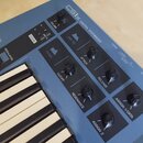 Modrý kontrolní syntetizátor s nápisem CSIX CONTROL SYNTHESIZER. Přístroj má klávesnici a několik ovládacích knoflíků s popisky VOLUME, ATTACK, RELEA…