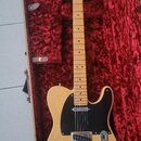 Elektrická kytara typu Telecaster v přírodním dřevěném odstínu s černým pickguardem, umístěná v otevřeném kufru s červenou výstelkou. Na hlavě hmatní…