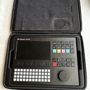Černý sampler Polyend Tracker v černém přepravním pouzdře. Zařízení má displej, mřížku bílých tlačítek a několik barevných ovládacích prvků s nápisy…