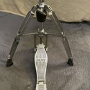 Stříbrný kovový stojan s pedálem, pravděpodobně hi-hat. Na šlapadle je vyrytý nápis MAPEX. Povrch kovových částí je lesklý, na spodní části nožek jso…
