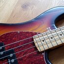 Detail elektrické basové kytary s tělem v přechodovém lakování od žluté po tmavě hnědou. Nástroj má červený mramorovaný pickguard a světle barevný hm…