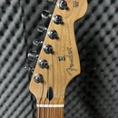 Fotografie zachycuje hlavu elektrokitary z přírodního dřeva s chromovanými ladičkami. Na dřevě je vidět nápis „Fender STRATOCASTER®“ a v horní části…