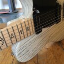 Elektrická kytara s bílým trupem z výrazně zrněného dřeva a světlým hnedým hmatníkem. Nástroj má černý pickguard, dva snímače typu humbucker a kovový…