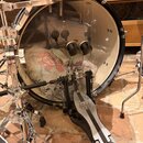 Fotografie zachycuje basový buben s průhledným předním koženem a přiložený pedál. Na pedálu je viditelný nápis MAPEX. Vnitřek bubnu je vyplněn textil…