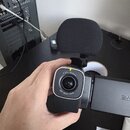Černá digitální kamera s připevněným externím mikrofonem v černé pěnové clone. Kolem objektivu je bílý nápis F 2.0 / 16.6mm a 160° WIDE LENS. Na prav…
