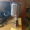 Stříbrný mikrofon s kovovou mřížkou v retro stylu, umístěný na stolním stojanu. Na přední straně mřížky je viditelné kulaté logo s písmenem „S“. Na s…