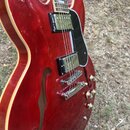 Červená polodubová elektrická kytara s krémovým lemováním a černým pickguardem. Nástroj má dva humbuckery s kovovými kryty a čtyři ovládací knoflíky.…