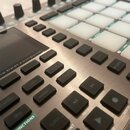 Detailní pohled na ovládací panel stříbrného MIDI kontroléru. Na zařízení jsou umístěna bílé podsvícené pady a černé plastové tlačítka s nápisy, mezi…