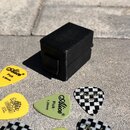 Černá plastová krabička s viditelnou texturou 3D tisku a zapadacím víkem. Kolem krabičky je rozprostřeno několik trsátel různých barev a značek. Na ž…