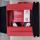 V červené krabici jsou umístěna červenobílá sluchátka. Na vložce je nápis FREE WARRANTY EXTENSION a KOSTENLOSE GARANTIEVERLÄNGERUNG. Vedle ní je štít…