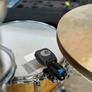 Černý mikrofon s mřížkou a logem Sennheiser v kruhu, upevněný na držáku k žlutému bubnu. Na těle mikrofonu je nápis e 604. Mikrofon je umístěn u bílé…