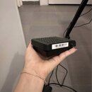 Černý, čtvercový box s mřížkou otvorů na horní straně. Na boku je nalepený bílý štítek s čárovým kódem a textem 0F4AACD58C84AAA. Zařízení je v ruce a…