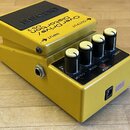Žlutý efekt pedál s černým ovládacím panelem a čtyřmi černými knoflíky s bílými značkami. Na horní části je nápis OverDrive/Distortion OS-2 a šipky O…