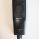 Černý kondenzátorový mikrofon s kovovou mřížkou v horní části. Na těle je bílý nápis maono a níže text Condenser Microphone. Povrch mikrofonu vykazuj…