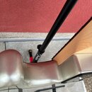 Detail stříbrného trupu elektrického nástroje s viditelným dřevěným hmatníkem. V pozadí je černý stojan a červená stěna. Na části stojanu je bílý náp…