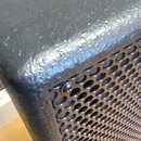 Detailní záběr na horní roh černého reproduktorového boxu s texturovaným povrchem. Přední strana je kryta kovovou mřížkou s drobnými kulatými otvory.…