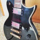 Černá elektrická kytara s lesklým povrchem a bílými lemovkami. Nástroj má dva humbuckery s kovovými kryty, zlatý kobylník a tři černé oválné ovládací…