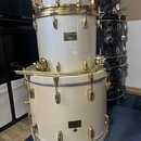 Tři bílé bubny s pozlaceným hardwarem naskládané na sebe. Na horním a prostředním bubnu je viditelné logo Pearl, přičemž na prostředním je nápis Mast…