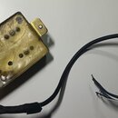 Elektrický snímač typu humbucker s mosazným nebo zlatým krytem. Povrch je výrazně znečištěný, s viditelnými skvrnami a oxidací. Z nástroje vede černý…