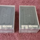 Dvě černé elektrické kytarové snímače typu humbucker, každá v průhledném plastovém obalu. Na spodní části obalů je bílý nápis METAL MACHINE. Snímače…