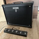 Černý kytarový kombozesilovač s logem BOSS a pod ním symbolem stylizovaného meče. Před zesilovačem je umístěn černý nožní spínač s nápisy BOOSTER, MI…