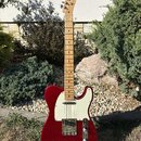 Elektrická kytara typu Telecaster v červené barvě s bílým pickguardem a světle hnědým javorovým hmatníkem. Na hlavě je viditelné logo Fender. Povrch…
