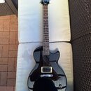 Černá elektrická kytara s tmavým hnedým hmatníkem a černým pickguardem. Na hlavě je bílý nápis Epiphone. Nástroj vykazuje známky běžného používání, p…