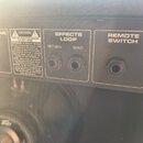 Detail zadní strany černého zesilovače s ovládacími prvky a varovnými štítky. Jsou zde vidět vstupy s popisky EFFECTS LOOP (RETURN, SEND) a REMOTE SW…