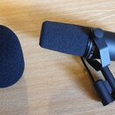 Černý kondenzátorový mikrofon v držáku s nasazenou pěnovou clonou. Vedle mikrofonu leží druhá, samostatná černá pěnová clona. Na těle mikrofonu je vi…