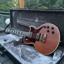 Elektrická kytara s hnědým trupem a tmavě červeným pickguardem v plastovém kufru s šedým polstrováním. Na hlavě hmatníku je viditelné logo Epiphone.…