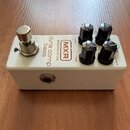 Bílý efekt pedál v kovovém krytu se čtyřmi černými oválnými knoflíky. Na horní straně jsou nápisy „dyna comp bass“, „BASSINNOVATIONS“ a logo „MXR“. U…