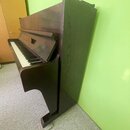 Tmavně hnědé digitální piano s dřevěnou texturou povrchu. Nástroj má klasický tvar s klaviaturou a integrovaným stojanem. Na viditelných částech nejs…