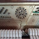 Detail vnitřku klavíru s kovovým rámem zlaté barvy a viditelnými strunami a kladívky. Na rámu je umístěn kulatý černobílý štít s nápisem ORIGINAL ROS…