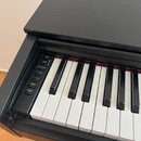 Černý digitální klavír s bílými a černými klávesami. V ovládací sekci vlevo jsou tlačítka s nápisy DEMO SONGS, GRAND PIANO, METRONOME, + R, - L, REC…