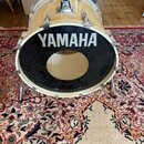 Světle béžový basový buben s černým předním koženem. Na koženě je bílý nápis YAMAHA a uprostřed je vyříznutý kruhový otvor. Nástroj má chromované lug…