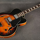 Polodupatá elektrická kytara s trupem v barevně sunburst (černá, oranžová a žlutá). Na hlavě hmatníku je bílé logo Ibanez. Nástroj má černý pickguard…