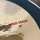 Detailní záběr na mosazný činel. Na povrchu je červeným písmem nápis WILD CRASH a pod ním označení 19". Povrch činelu vykazuje kování v podobě drobný…
