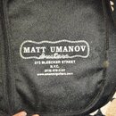 Černý textilní obal s bílým potiskem. Uprostřed je logo v rámečku s nápisem MATT UMANOV Guitars. Pod ním je uveden text 273 BLEECKER STREET N.Y.C. (2…