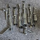 Sada kovových komponentů bicích pedálů v stříbrné barvě, rozložených na šedém koberci. Jsou zde vidět různé části mechanismů, včetně dvou nožních zák…