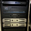 Černý transportní rack s několika zařízeními. Z horní části jsou vidět přístroje s nápisy „DISTRIBUTIVIG“, „QSC AUDIO Professional Power Amplifier US…