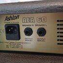 Zadní strana zesilovače v hnědém koženěném potahu s kovovým panelem. Na panelu je nápis Ashton, Designed in Australia a model AEA 60. Jsou zde dva vý…
