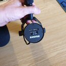 Fotografie zachycuje spodní část černého mikrofonního držáku s logem SHURE. V horní části jsou vidět dva USB porty. Povrch je černý, matný a drží jej…